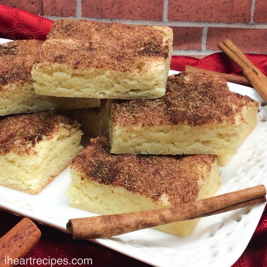 Chewy Snickerdoodles Bars stacked high on a lacy white platter and garnished with fragrant cinnamon sticks. 