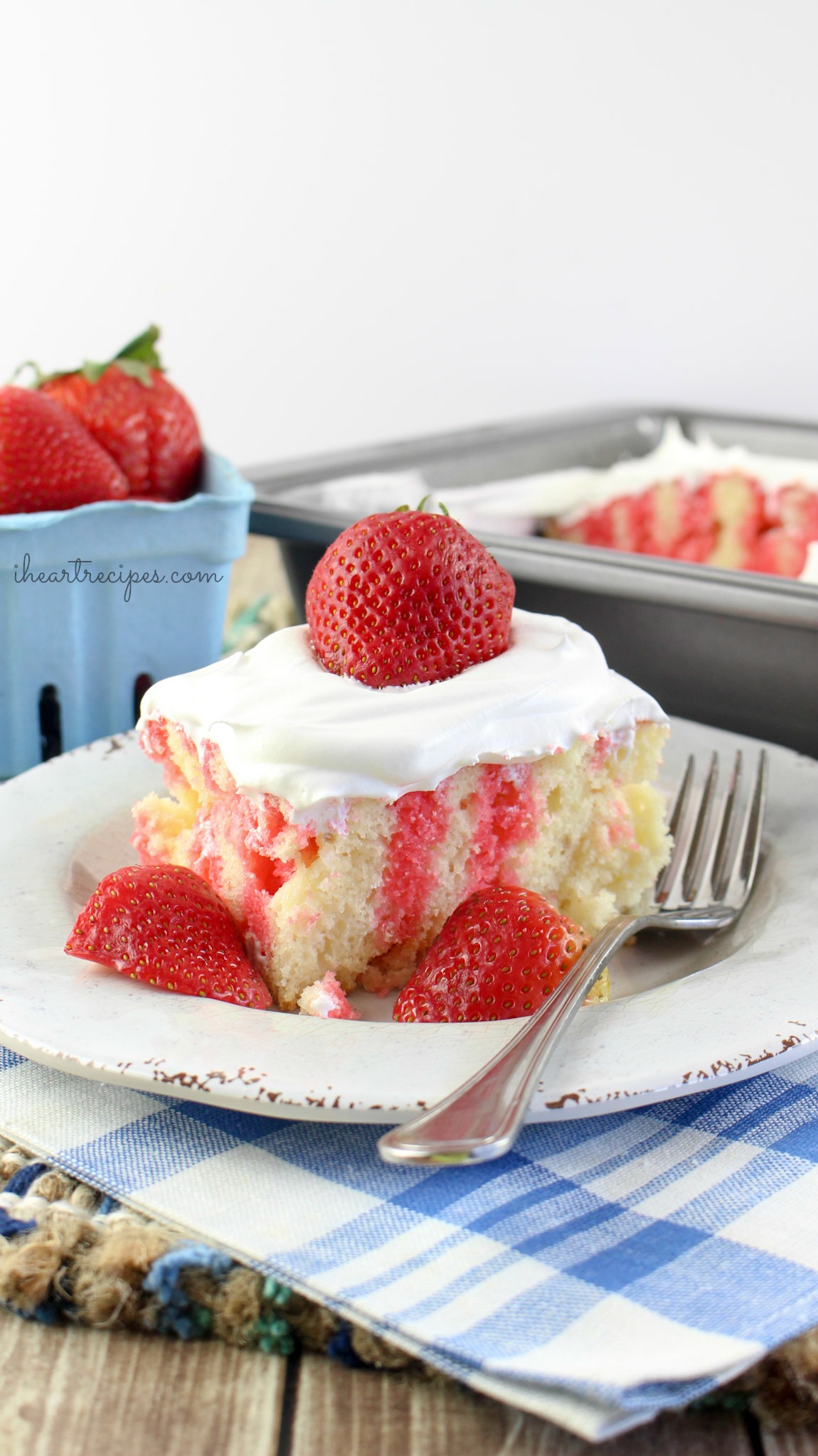 A moist strawberry jello poke cake topped with creamy whipped cream and fresh strawberries on a rustic white plate ready to be served. A delicious dessert!