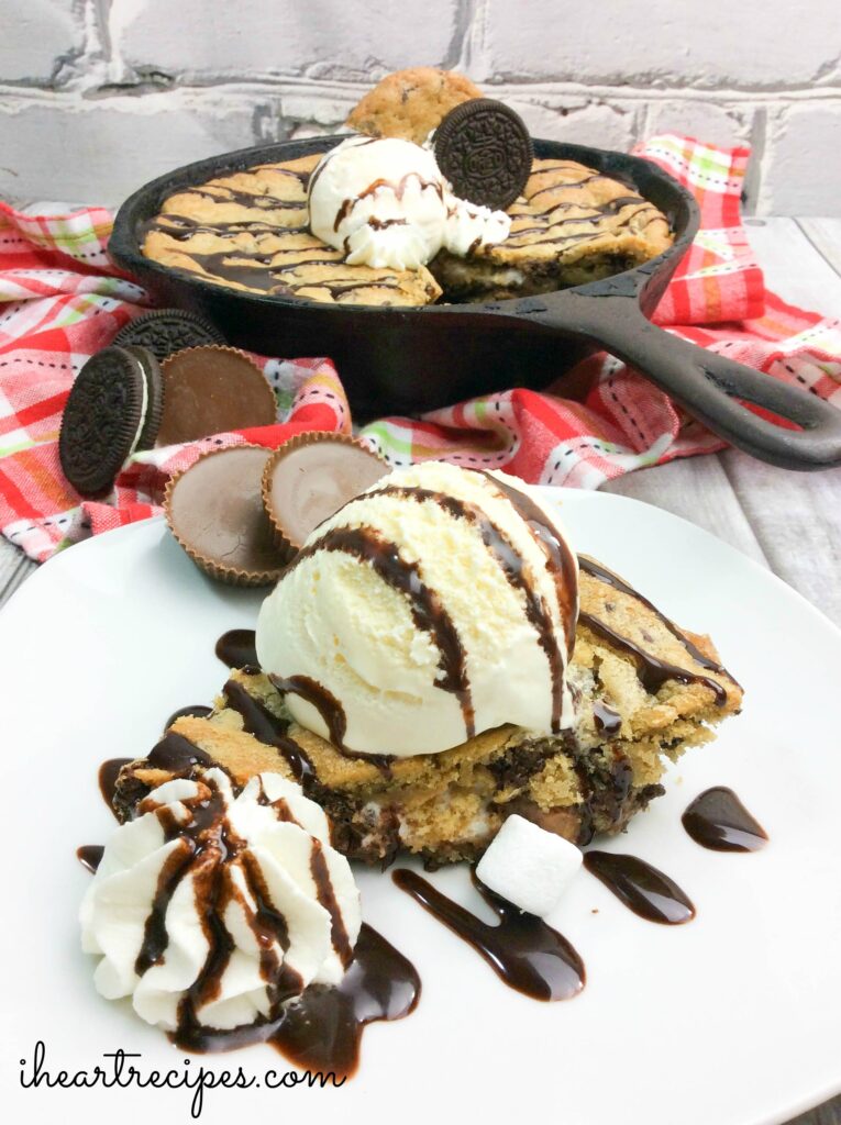A slice of skillet cookie served on a plate with ice cream, whipped cream, and chocolate sauce, and the full skillet cookie in a cast iron pan is seen in the background.