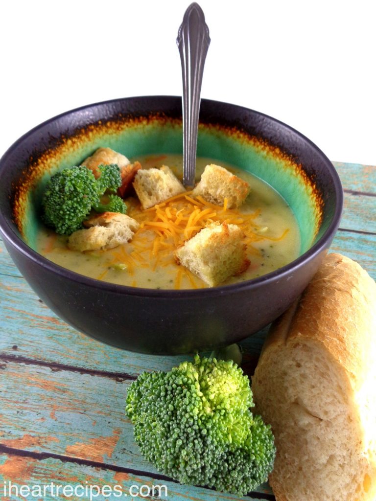 A bowl of homemade cheddar broccoli soup topped with melted cheese and crunchy croutons. The bowl is set on a rustic wooden table next to fresh bread and broccoli. This meal will warm you right up!