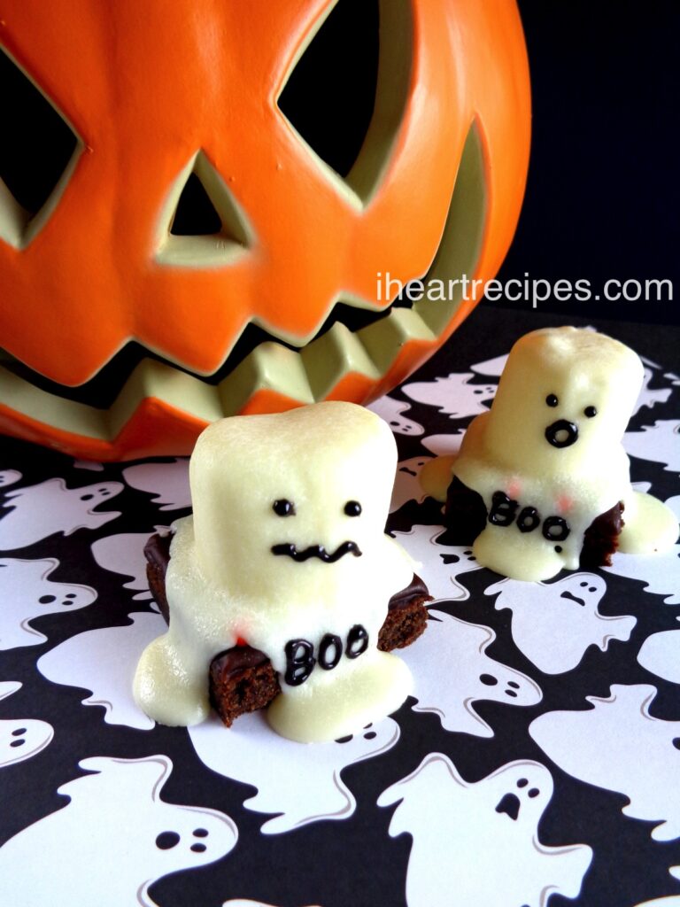 Two ghost brownies alongside a large orange pumpkin all set on a decorative black and white ghost tablecloth. 