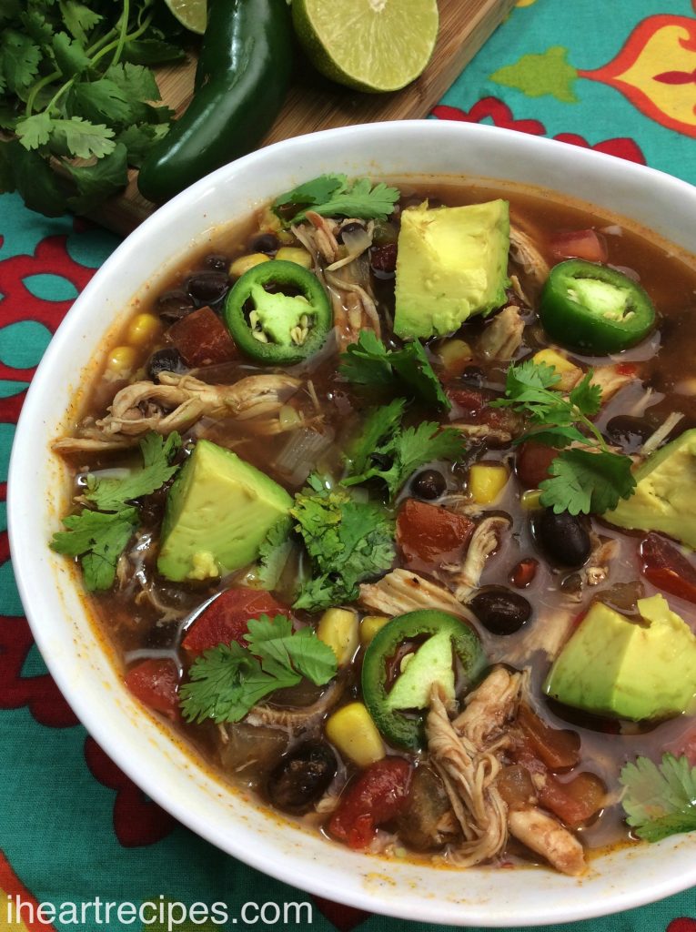 A white bowl filled to the brim with chunks of tender chicken and beans, bright veggies, and avocado chunks in a savory broth garnished with fresh cilantro. 