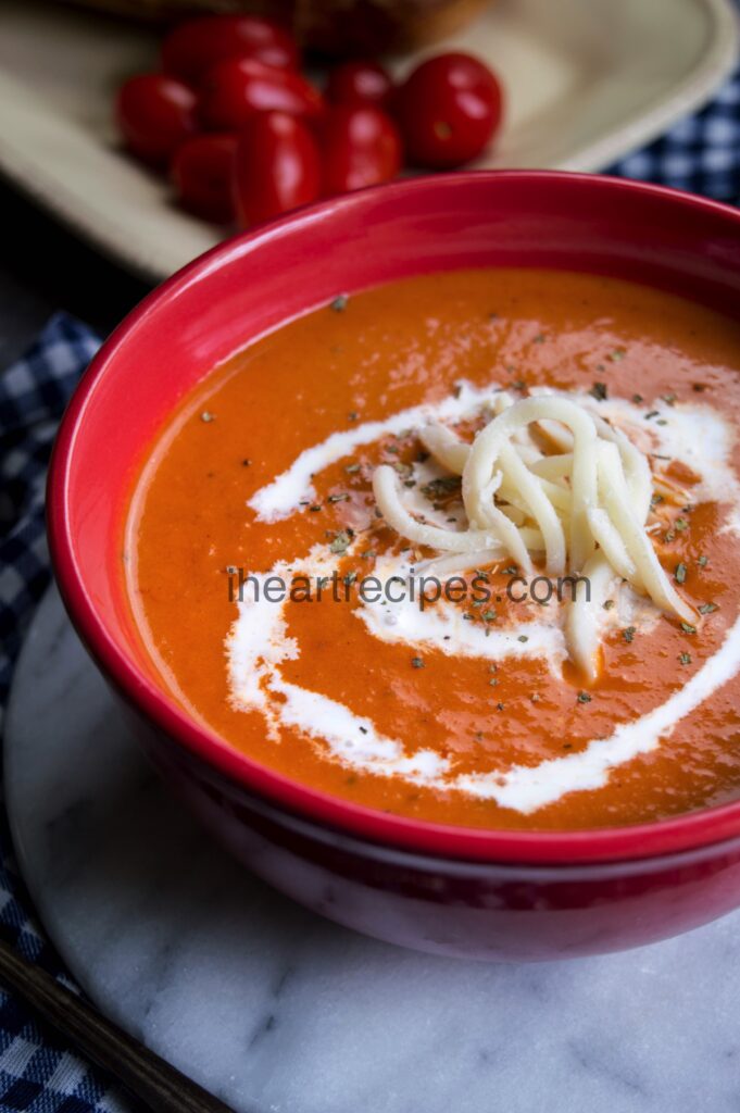 Creamy and delicious tomato soup  fills a red bowl on top of a granite cutting board. A bunch of small red tomatoes sit in the background. 