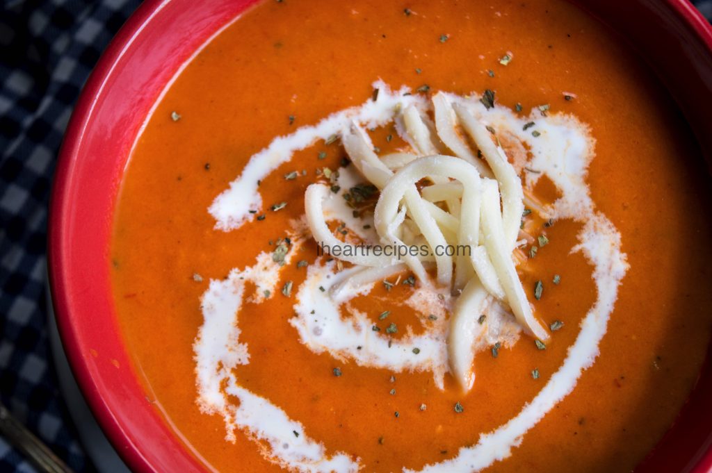 A close up image of a bowl of fresh tomato soup topped with shredded white cheese, a swirl of sour cream and herbs. 