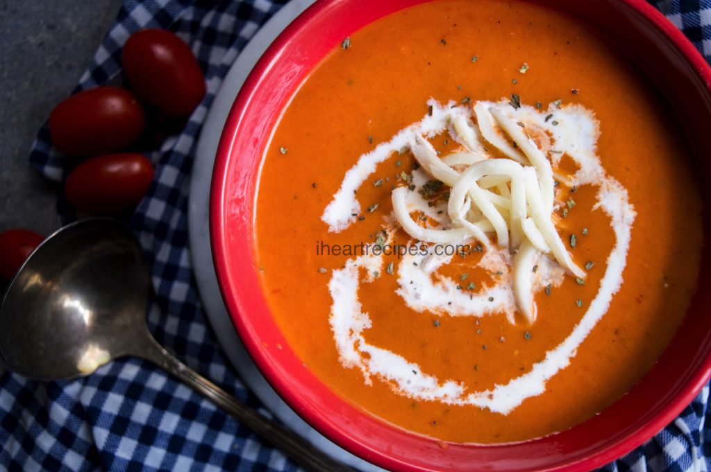 A red bowl filled with fresh tomato soup and topped with a swirl of sour cream, cheese and seasoning. The perfect comfort meal on cold winter days.