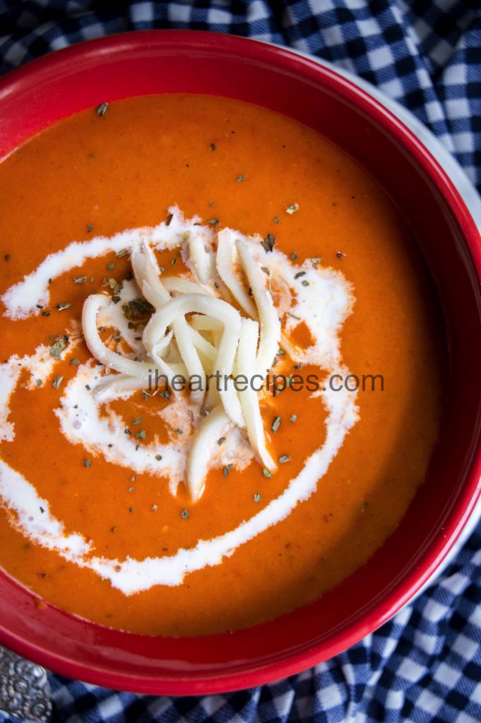 An overhead image of a red bowl filled with fresh tomato soup garnished with white cheese, a swirl of sour cream and seasonings. 