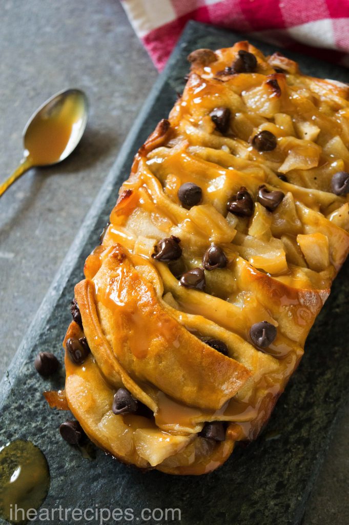 A close-up of caramel apple pull apart bread showcasing the layers of golden dough with bits of sweet apple and melted chocolate chips tucked inside. It is served on a grey stone serving platter. A silver spoon coated in caramel sauce lay nearby.   