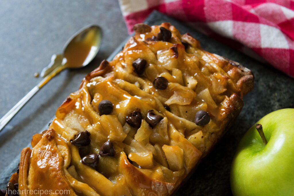 A close-up of gooey Caramel Apple Pull Apart Bread topped with sweet chocolate chips set on a marble serving platter. A green apple and silver spoon coated in caramel sauce lay nearby. 