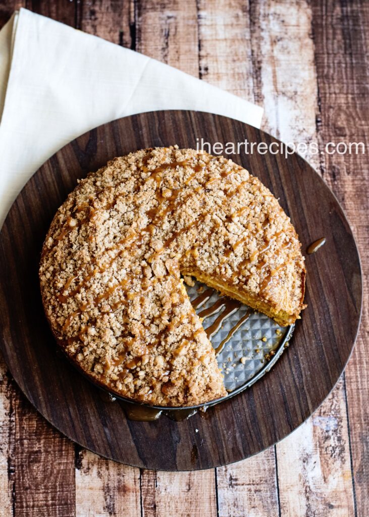 A delicious caramel pumpkin coffee cake with a golden crumble topping drizzled with sweet caramel sits on a rustic wood serving platter. A slice is removed.