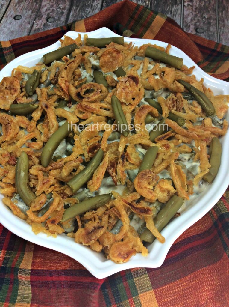 A white casserole dish filled with freshly baked green bean casserole sits on a red, green, and yellow plaid linen cloth. French-style cut green beans and golden-brown crispy fried onions top the creamy casserole.