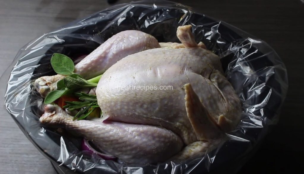 A whole raw turkey sits in a plastic bag-lined slow cooker. Leaves of fresh sage and other aromatics are stuffed into the turkey cavity.