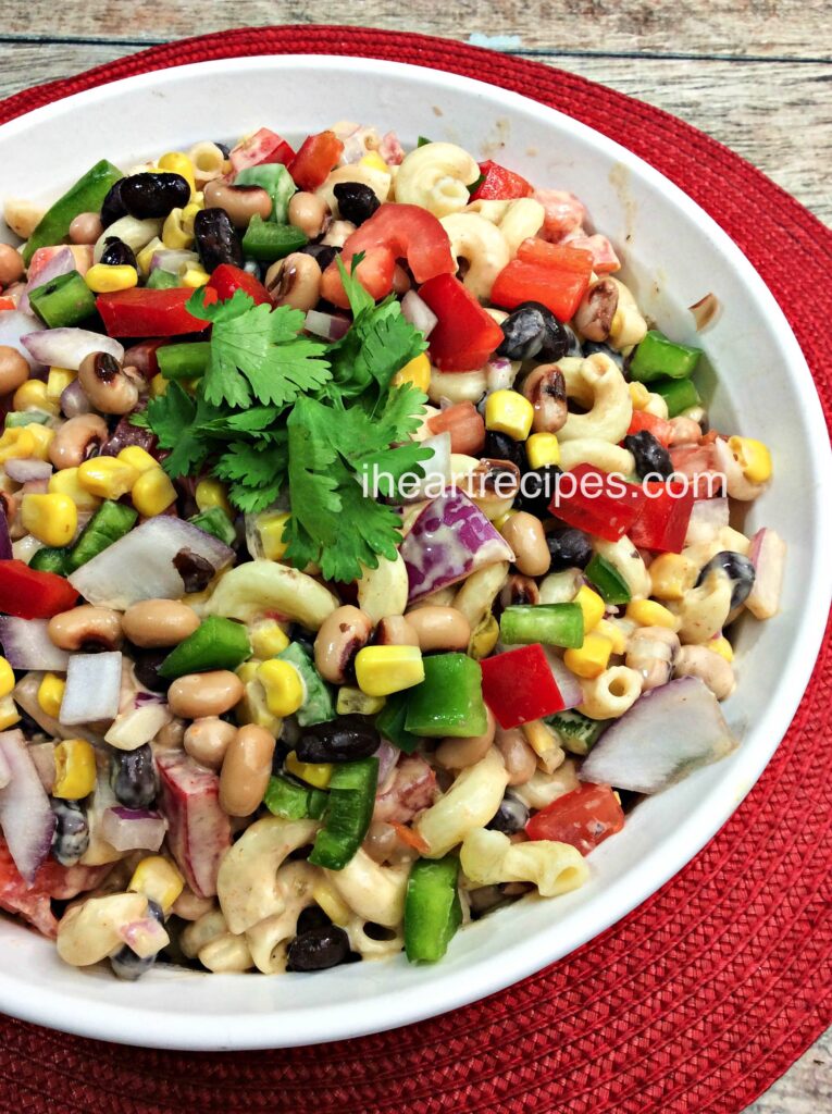 This beautiful and satisfying Texas Caviar Macaroni Salad is served in a white serving dish on top of a vibrant red woven placemat and placed on a rustic wooden table. 