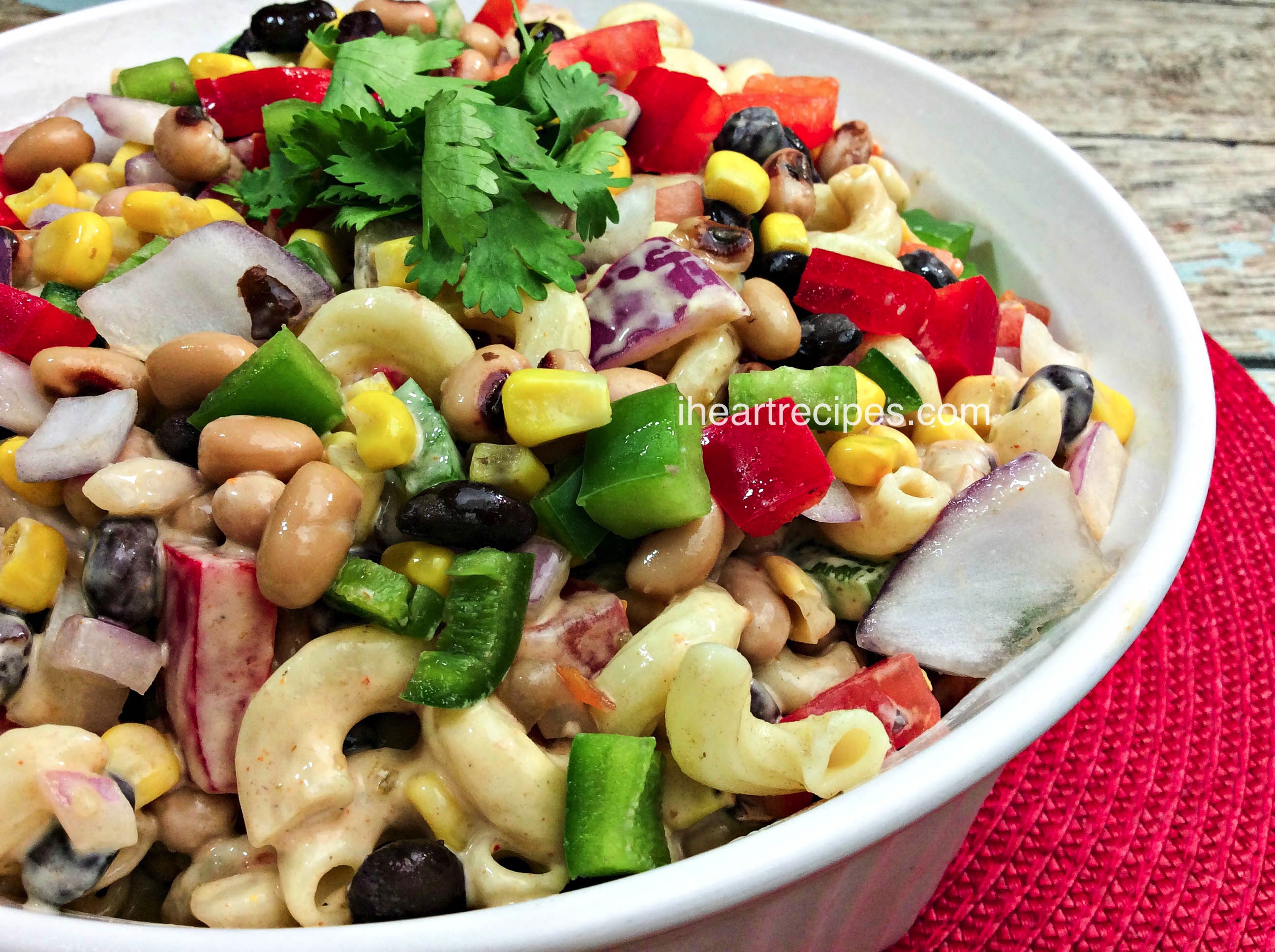 Flavorful and colorful Texas Caviar Macaroni Salad served in a classic white serving dish on top of a red woven placemat and placed on a rustic wooden table. It is a refreshing summer salad with a kick of heat!