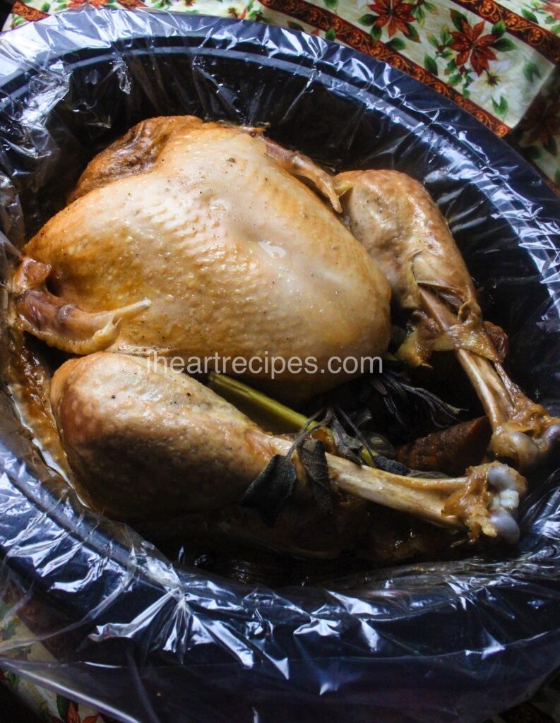 A whole turkey sits in a slow cooker, lined with a plastic slow cooker liner. The turkey is perfectly slow-cooked, the golden-brown skin and tender meat pulling back from the turkey leg bones.