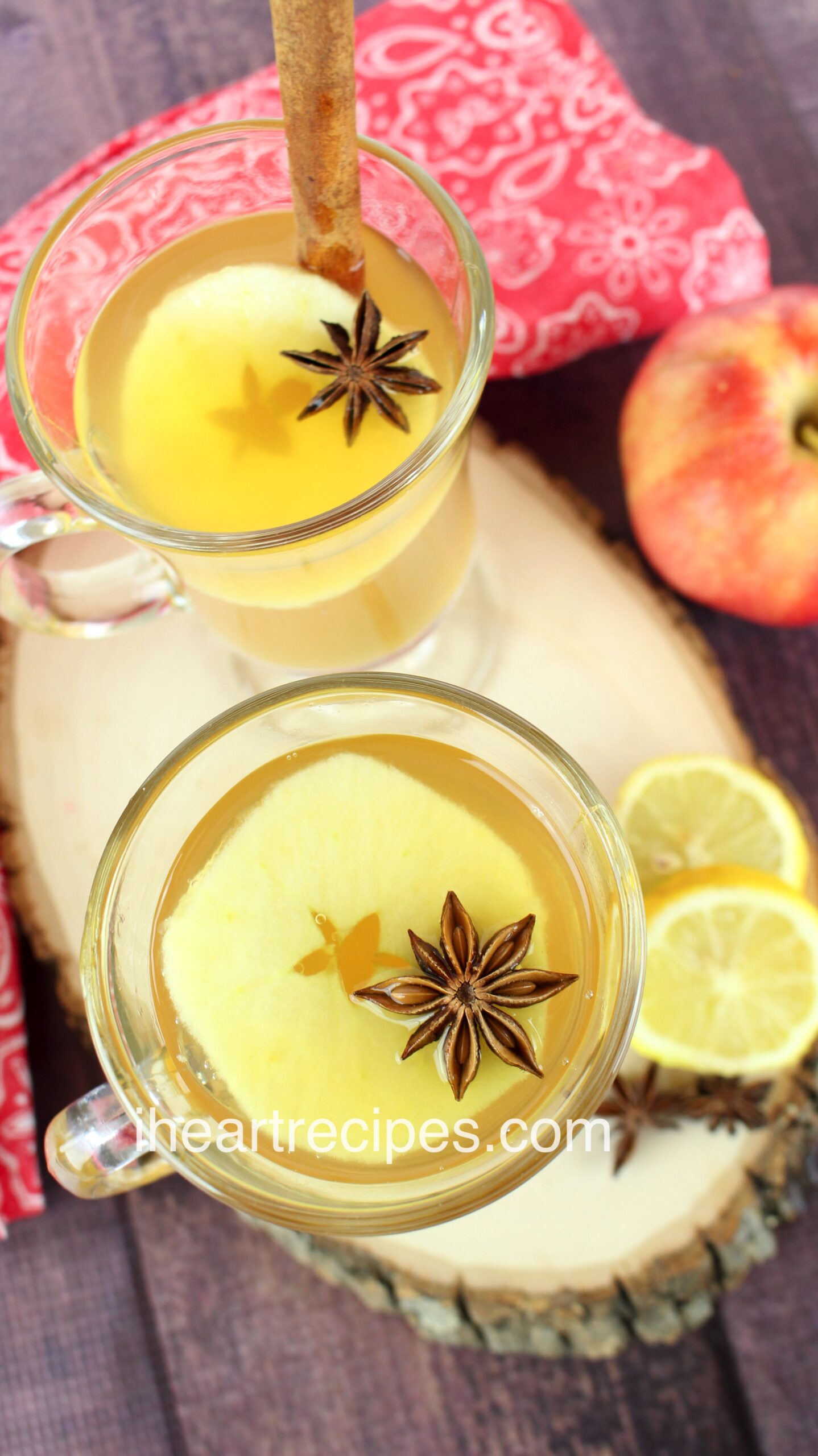 Two glasses of old fashioned hot toddy cocktail sit on a large wood platter. Each glass is garnished with apples slices, star anise, and a cinnamon stick.