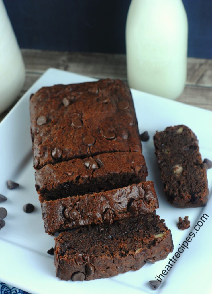 Moist Chocolate Banana Bread slices on a square white plate sprinkled with chocolate chips. A carafe of creamy milk is nearby. What a sweet treat!