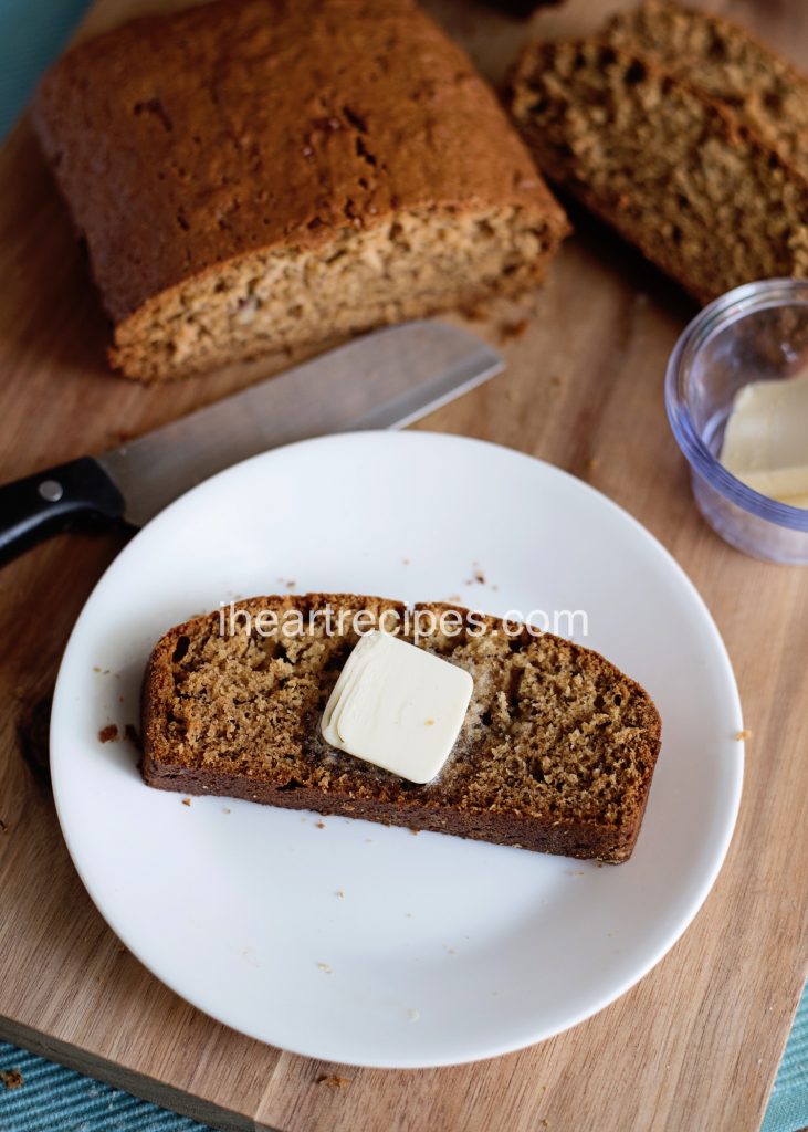 A slice of fresh, warm and moist banana bread served with a pat of butter. The remainder of the loaf is in the background alongside a knife and a bowl of butter. 