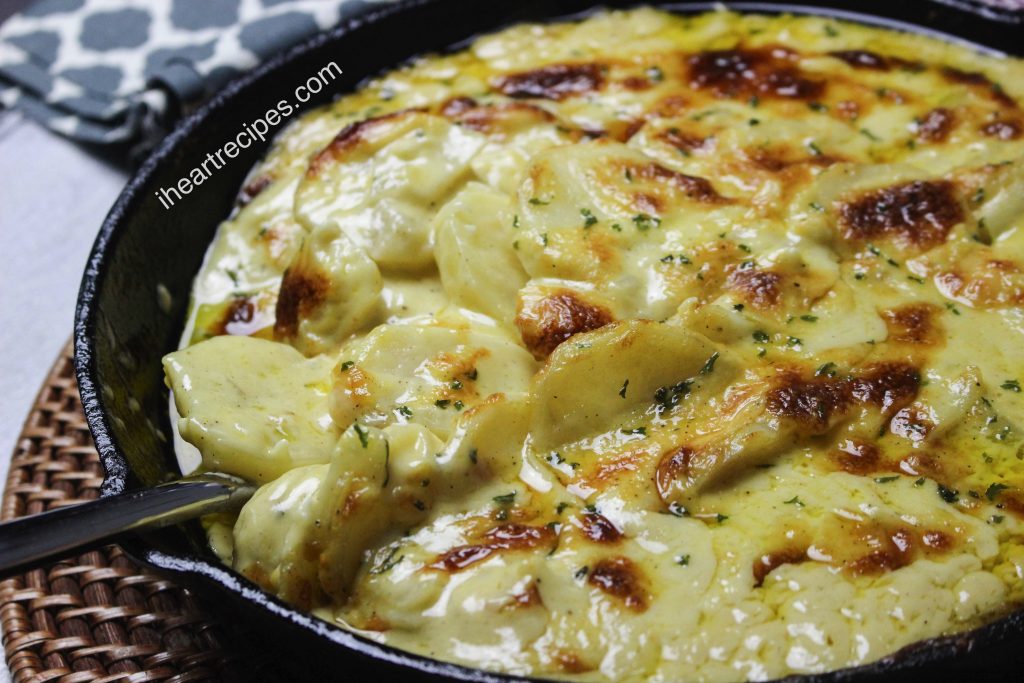 Golden, creamy and cheesy scalloped potatoes served in a skillet with a silver spoon. The skillet rests on a wicker placemat. 