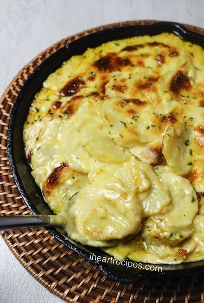 Creamy cheese and tender potatoes garnished with parsley flakes in a skillet. A silver spoon rests in the dish. 