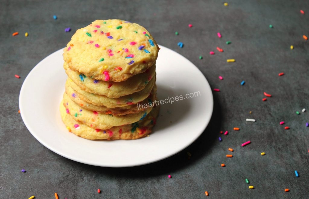 Cake mix confetti cookies piled neatly on a round white plate. The grey countertop is covered in rainbow sprinkles. 