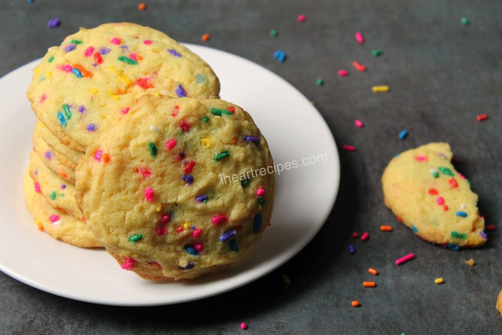 A stack of colorful cake mix confetti cookies on a white plate. There are colorful sprinkles and a half-eaten cookie on the counter. 