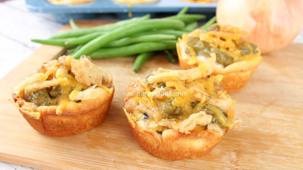 Three mini Green Bean Casserole Biscuit Cups on a wood cutting board. Fresh green beans and an onion are in the background. 