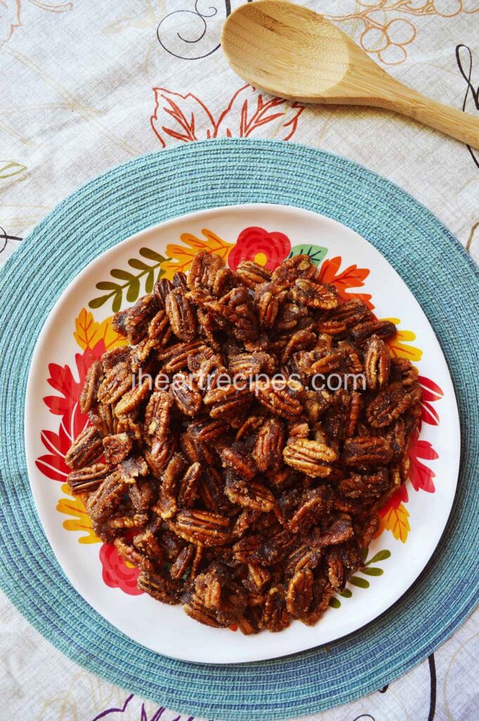 An overhead image of a pile of crunchy candied pecans on a festive fall plate set on a light blue placemat. 