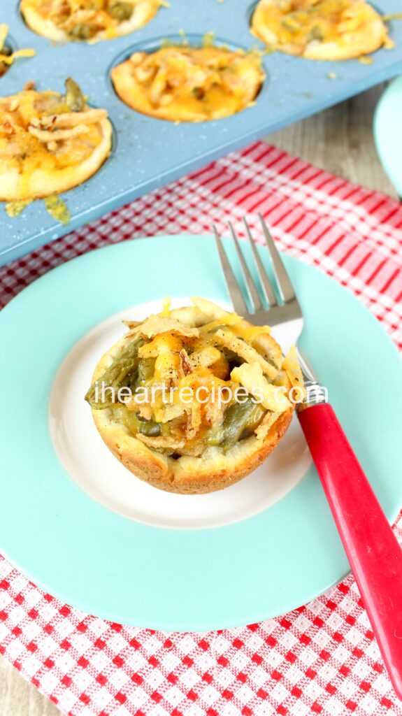 A single biscuit cup and a pink and silver fork on a blue plate. There is a muffin tin full of Green Bean Casserole Biscuit Cups in the background. 