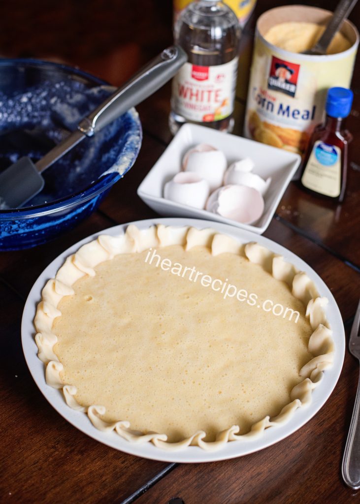 A white 9-inch pie pan filled with a pie crust and liquid, unbaked chess pie filling. The ingredients for chess pie sit behind the pie pan—eggs, white vinegar, cornmeal, and vanilla extract.