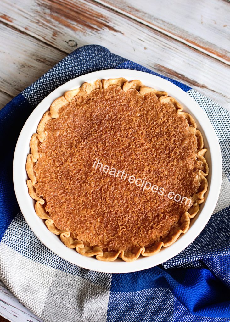 An old-fashioned chess pie baked in a white, round ceramic pie dish. The chess pie has a scalloped edge and a golden-brown top crust.