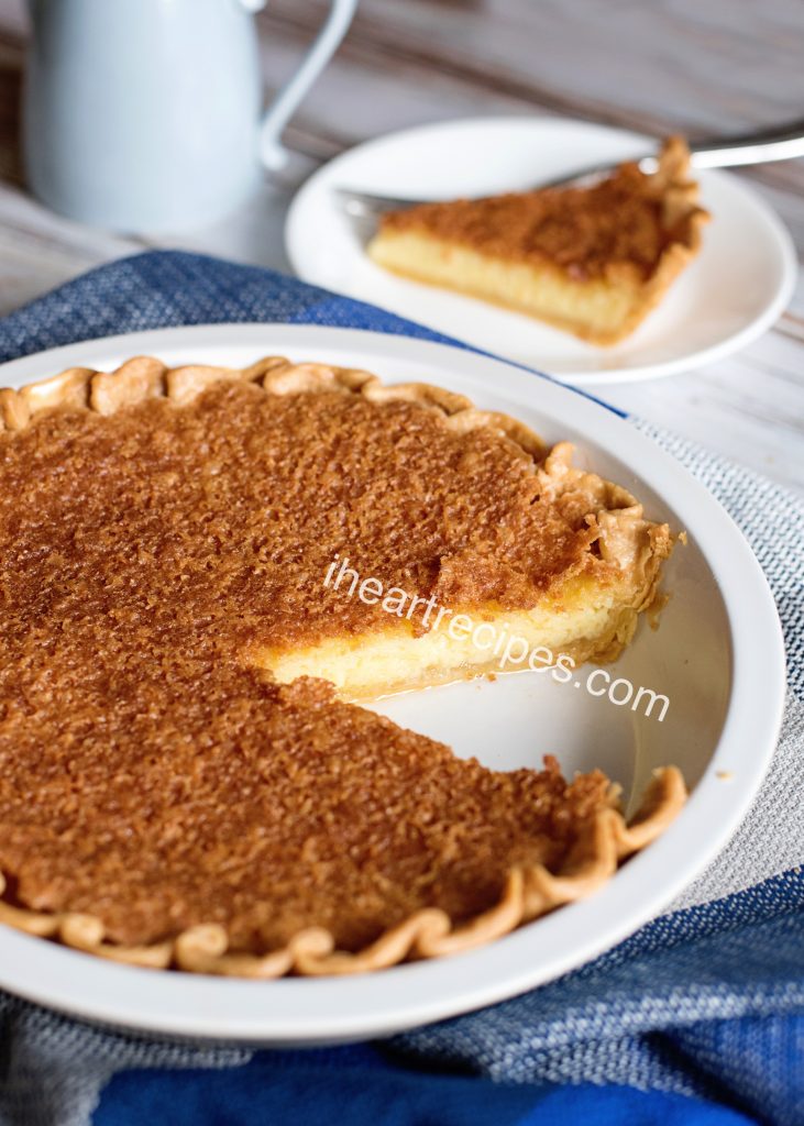 A homemade chess pie in a white ceramic pie dish, with one single slice cut out. The inside texture of the chess pie is creamy and custard-like, with a golden-brown top crust.