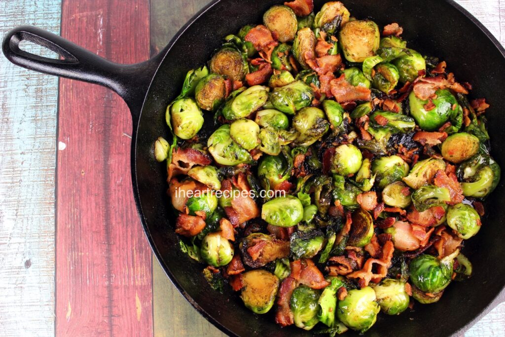 A cast iron skillet filled with roasted Brussels Sprouts topped with a sweet balsamic vinegar dressing and bacon bits. The skillet is set on a wood plank table. 