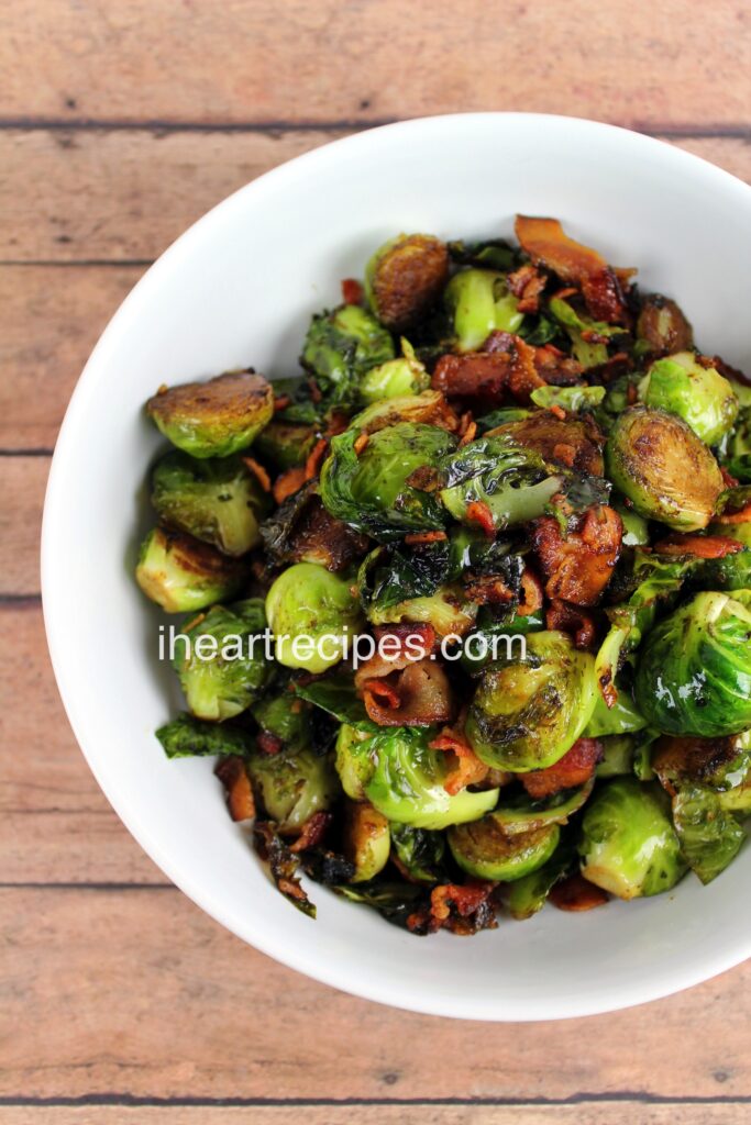 Roasted Brussels Sprouts tossed with balsamic vinegar and bacon pieces in a white serving bowl atop a wooden table. 