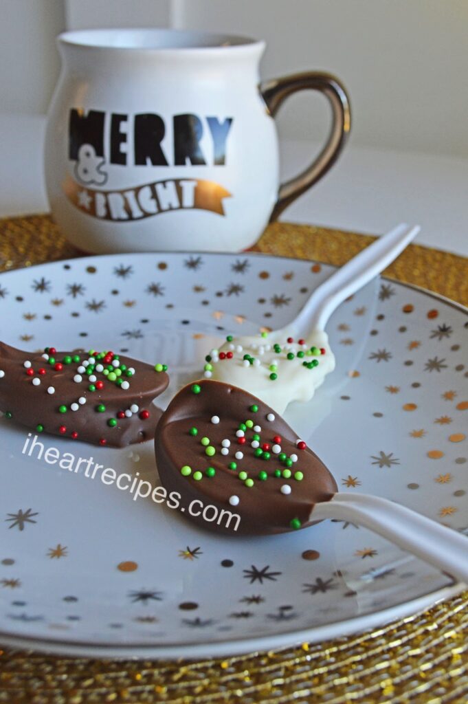 Three Hot Chocolate Spoons resting on a festive white, gold and silver plate. A white, black and gold mug in the background has the words Merry & Bright written on it. 