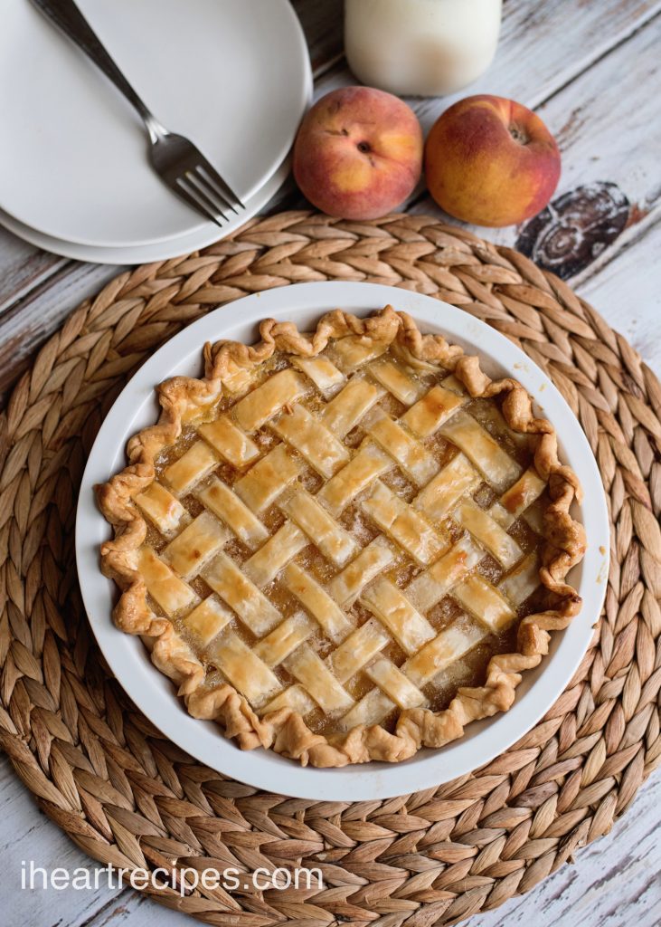 A homemade peach pie with a golden-brown lattice crust in a white ceramic pie dish sits on a woven placemat. White plates, forks, two fresh peaches, and a glass of milk sit next to the peach pie.