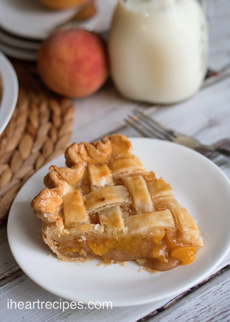 A slice of homemade peach pie on a white plate. The piece of pie has a golden-brown lattice pie crust and a perfectly-set peach pie filling