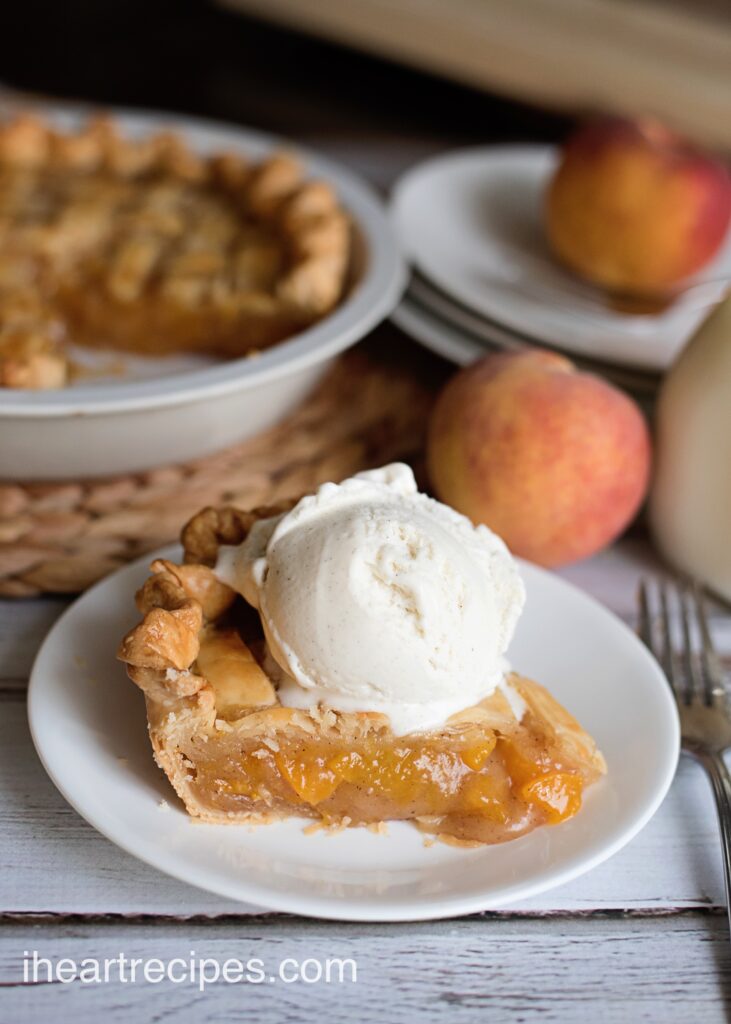 A single slice of peach pie topped with a scoop of vanilla ice cream on a white plate. Fresh peaches and a white pie dish with a whole peach pie are in the background.