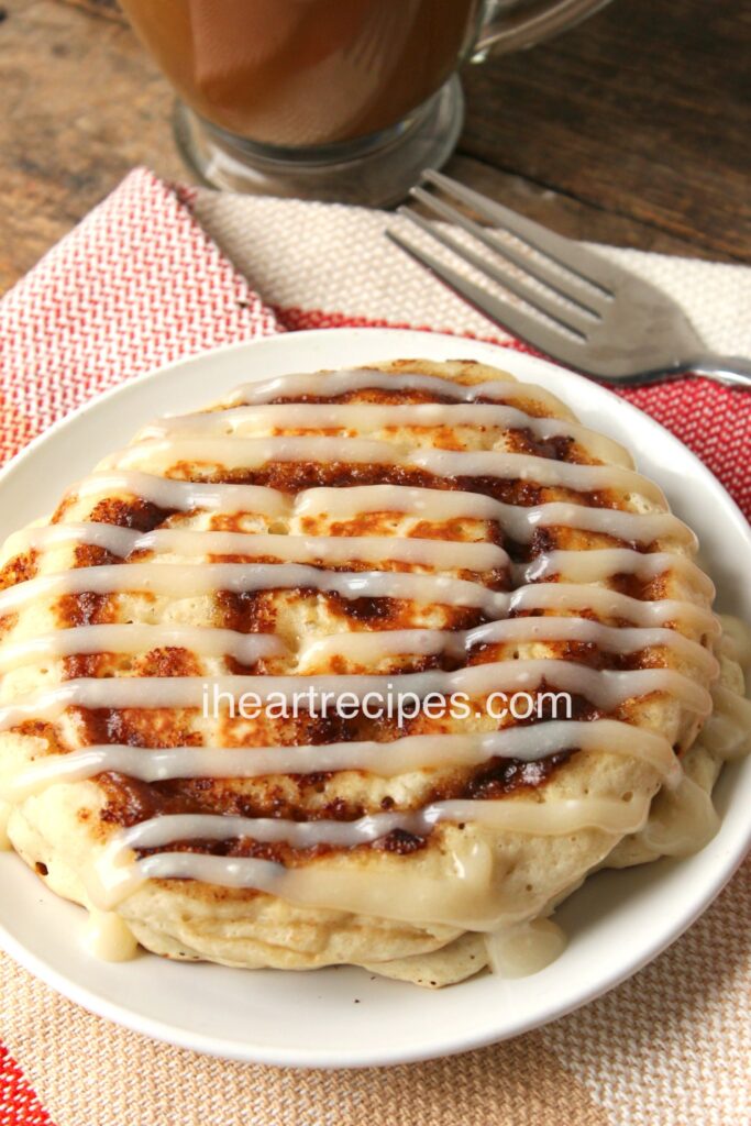 From-scratch cinnamon roll pancakes served on a plate. These pancakes have a sweet cinnamon sugar swirl and a drizzle of homemade cream cheese icing on top.