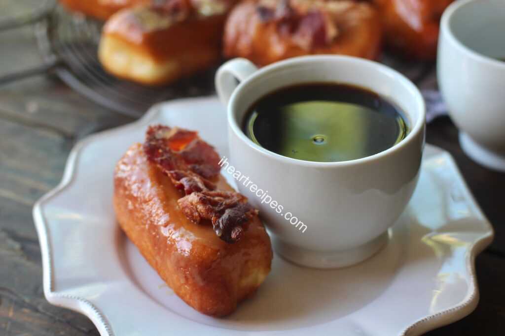 A sweet maple bacon donut next to a small cup of black coffee, both set on a saucer. A tray of donuts is in the background. 