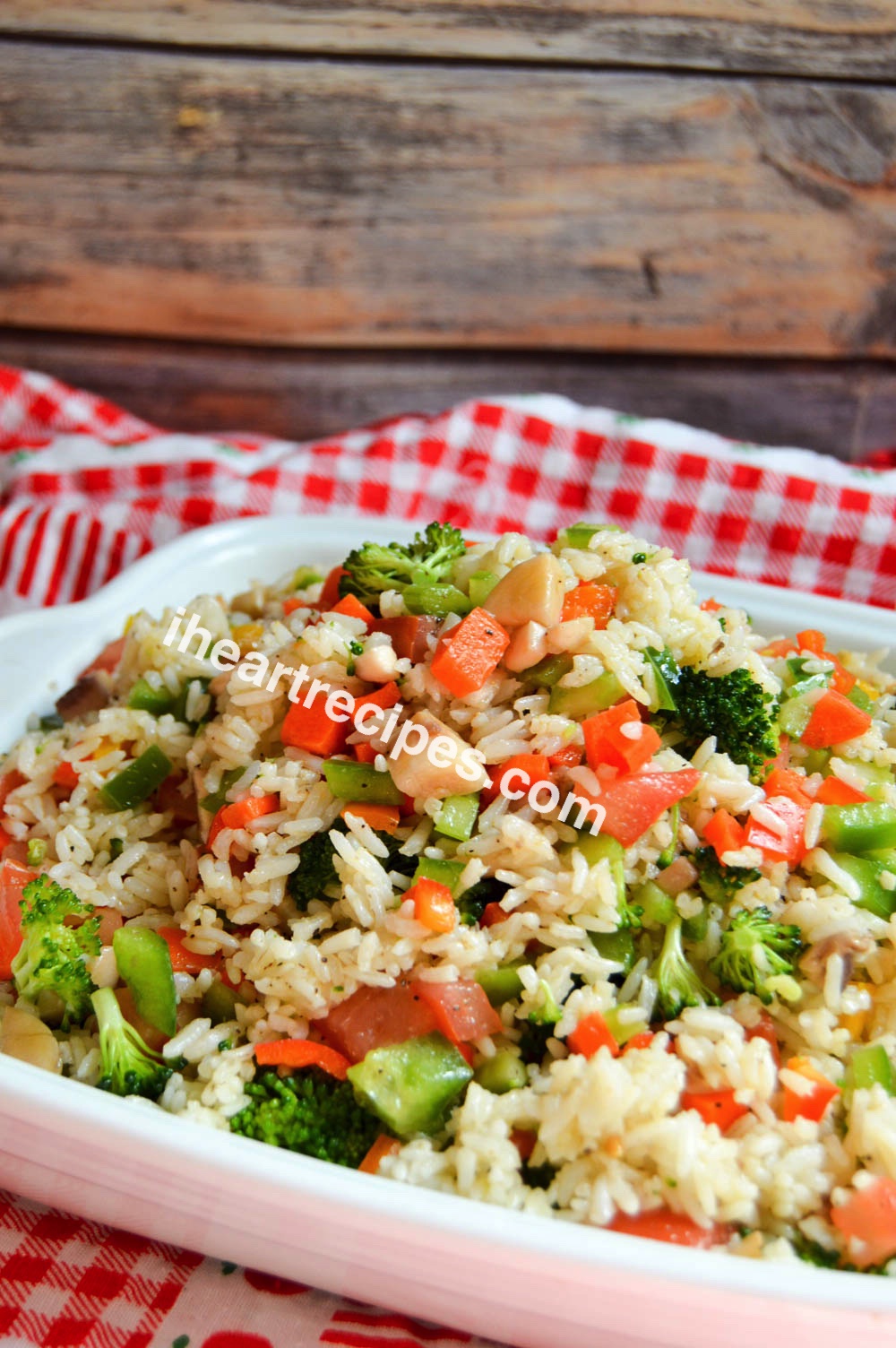A casserole dish filled with rice salad sits on a red and white checkered tablecloth. The rice salad is a mixture of white rice and broccoli, diced bell peppers, celery, and carrots.