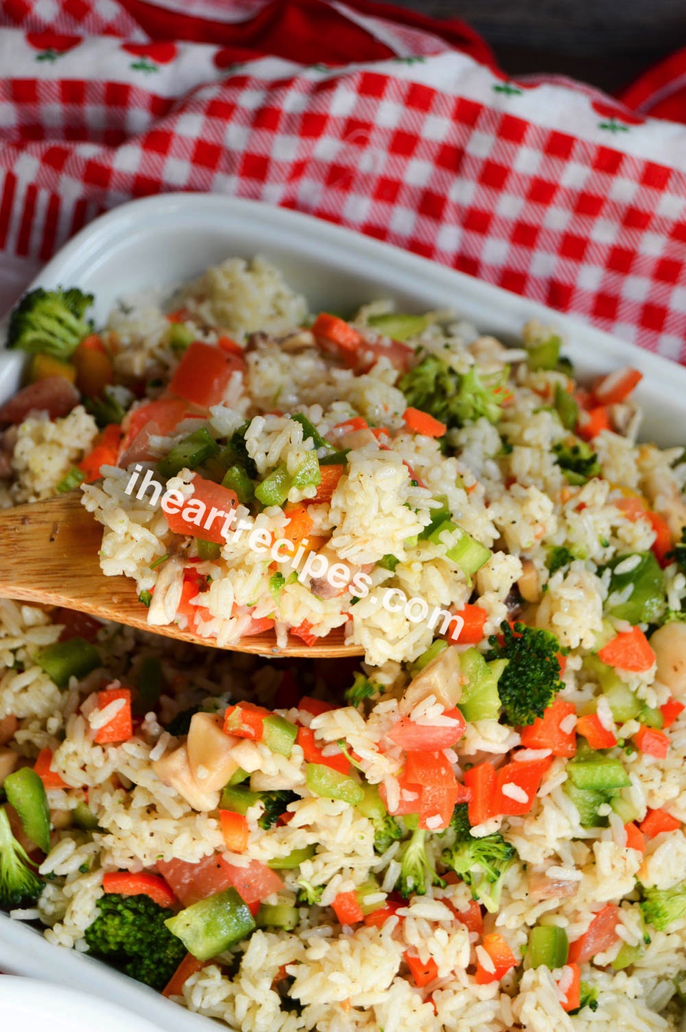 A wooden spoon scoops a serving of cold rice and vegetable salad from a white casserole dish.