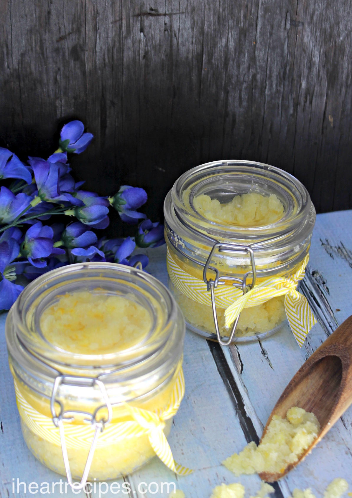 Golden-yellow lemon body scrub in clear glass jars wrapped in a yellow and white ribbon sitting on a rustic wooden table. It's like sunshine in a jar!