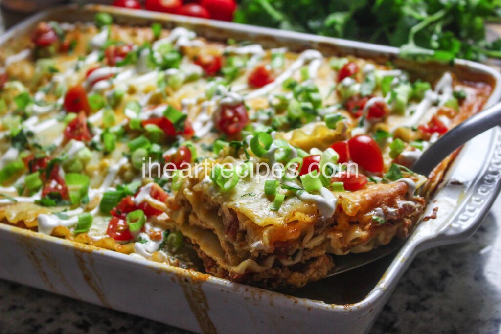 A slice of layered taco lasagna with ground turkey is cut from a casserole dish. This taco lasagna has layers of seasoned taco meat, cheese, and lasagna noodles, topped with veggies and sour cream.