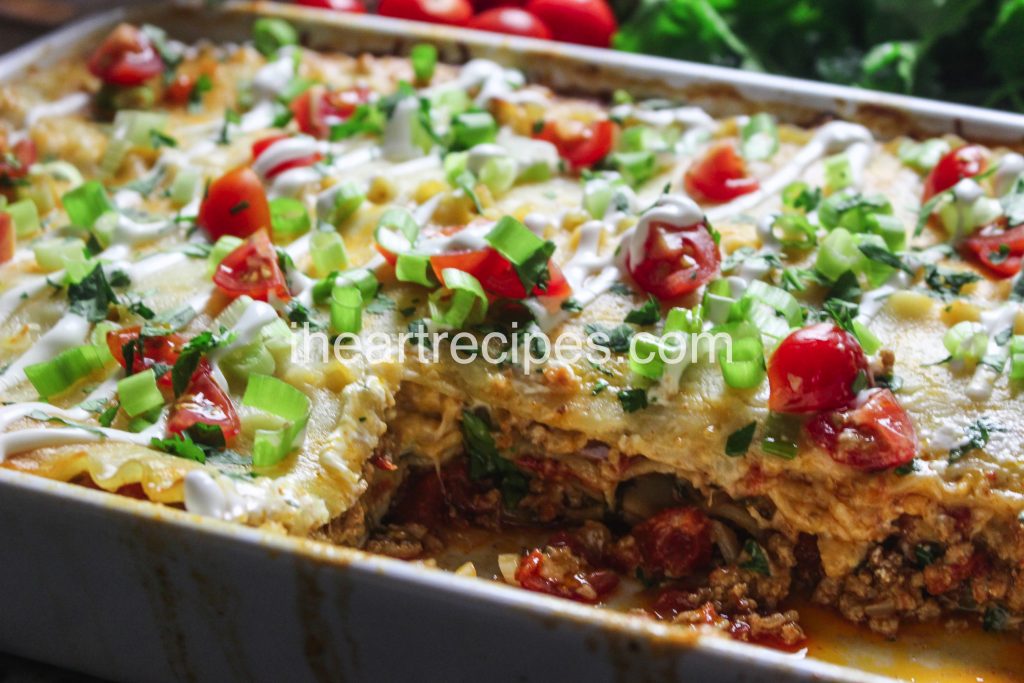 A casserole dish with a few slices of taco lasagna cut out, revealing melty layers of seasoned ground turkey, cheese, lasagna noodles, and topped with green onions, tomatoes, cilantro, and sour cream.