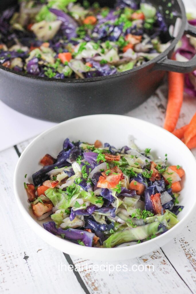 Colorful cabbage stir fry with bacon in a white bowl garnished with fresh parsley. A dark grey pan filled with stir fry is nearby. 