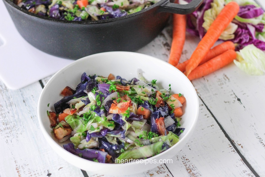 A white bowl filled with green and red cabbage, orange carrots and bacon.