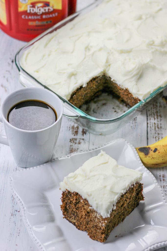 A slice of hummingbird cake on a square white plate served next to a white coffee mug filled with Folgers coffee. A baking dish with the remainder of the cake sits nearby as well as a ripe banana. A container of Folgers coffee is in the background. 