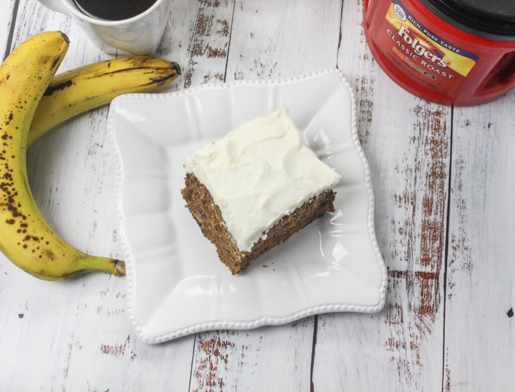 A square of Old fashioned hummingbird cake served on a white plate alongside a cup of Folgers coffee. A container of Folgers coffee as well as two ripe bananas sit nearby.