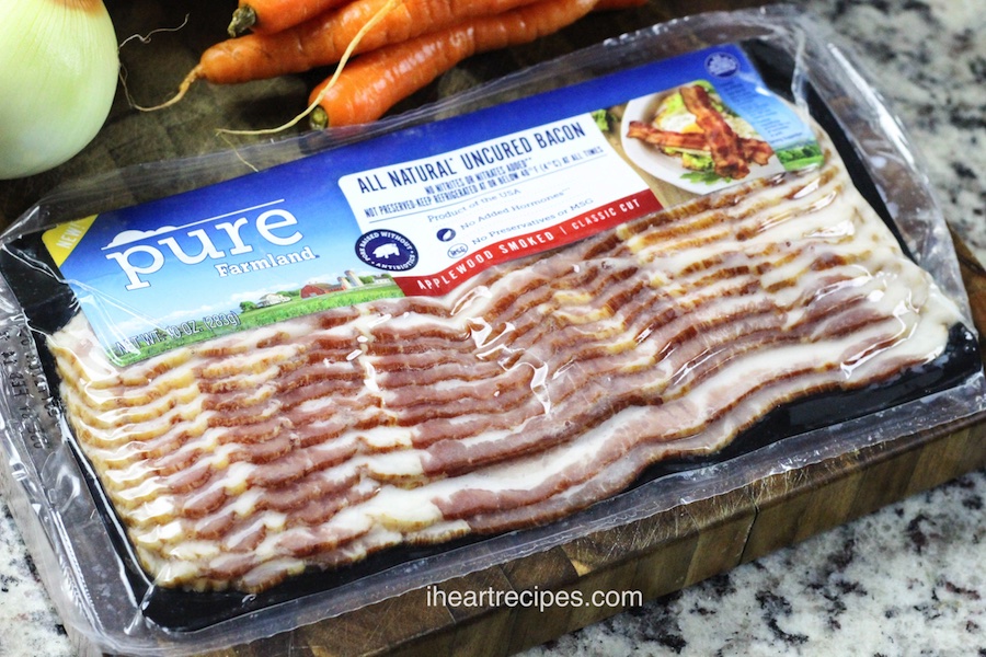 A package of Pure Farmland all natural uncured bacon on a wooden cutting board. An onion and carrots lay nearby. 