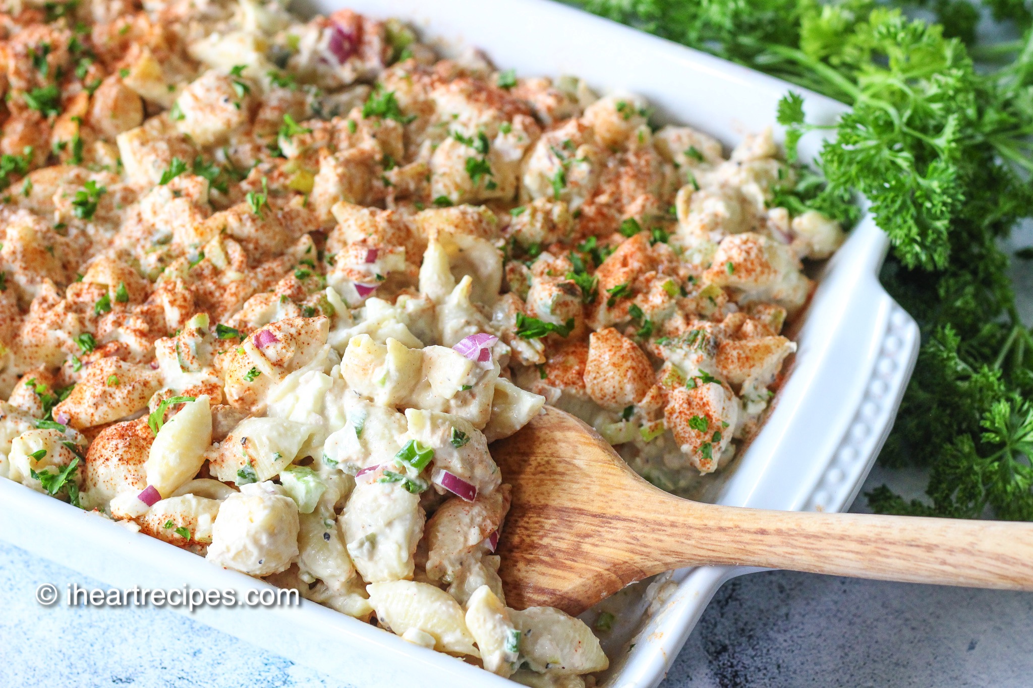 An overhead image of a casserole dish filled with creamy tuna macaroni salad. The pasta salad is topped with paprika and served with a wooden serving spoon.